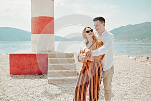 A couple stands near a lighthouse on the beach against the backdrop of the sea and mountains. Vacation love, honeymoon, travel.