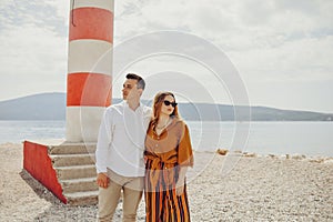 A couple stands near a lighthouse on the beach against the backdrop of the sea and mountains. Vacation love, honeymoon, travel.
