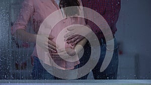 Couple stroking pregnant woman tummy behind rainy window, baby expectation