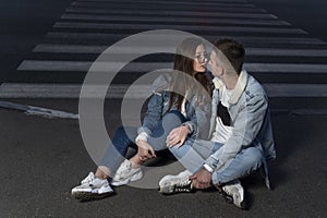 Couple of students in love are sitting on road in the late evening. Young girl and a guy on a date. First kiss