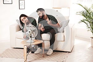 Couple suffering from heat in front of fan at home. Summer season