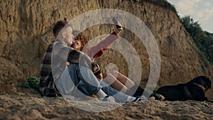 Couple taking selfie photo at sunset beach on smartphone. Happy lovers with dog