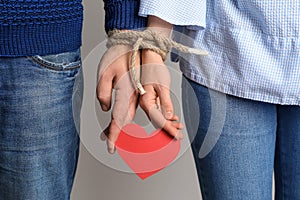 Couple with tied together hands holding paper heart on light background. Concept of addiction