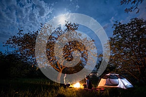 Couple tourists sitting at a campfire near tent under trees and night sky with the moon. Night camping