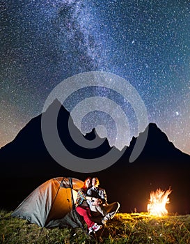 Couple tourists sitting near tent and campfire, looking to the shines starry sky and Milky way in the camping at night