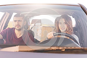 Couple travelers reach destination at car: beautiful smiling woman at wheel teaches to drive, her husband sits at front seat, cont