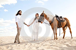 Couple of travellers holding hands, walking through desert on horse