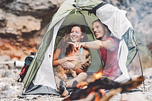 Couple of trekker sitting in the tent with their pet - Happy man and woman having fun in vacation camping around rock mountain