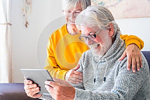 Couple of two cheerful and happy mature and old people seniors using tablet and having fun sitting on the sofa at home together.