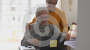 Couple of two happy seniors cooking together in the kitchen learning and following a video tutorial of cook in the laptop -