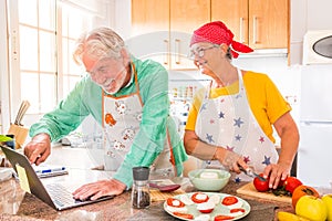 Couple of two happy seniors cooking together in the kitchen learning and following a video tutorial of cook in the laptop -