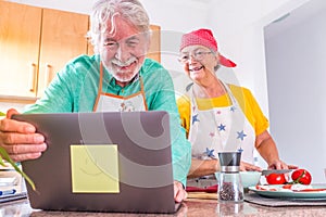 Couple of two happy seniors cooking together in the kitchen learning and following a video tutorial of cook in the laptop -