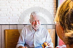 Couple of two seniors eating and drinking in a restaurant of fast food together - mature man holding an hamburger and prepared to