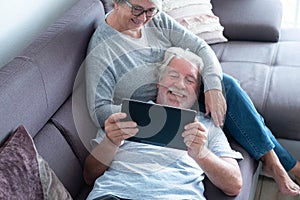 Couple of two seniors or mature people at home having fun together looking at the same tablet - pensioner lying on the sofa with