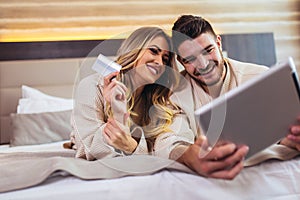 Couple using digital tablet and credit card while lying together in bed in hotel room