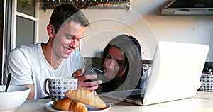 Couple using mobile phone and laptop while having breakfast