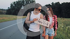 Couple on vacation hitchhiking along road and holding New York sign. Summer time. Hitchhiking, tourists, adventures