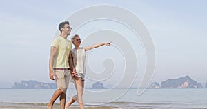 Couple Walking On Beach Holding Hands Talking, Young Man And Woman Point Finger, Tourists On Vacation