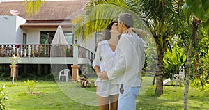 Couple Walking Embracing In Tropical Garden Near Villa House, Happy Man And Woman Kissing Outdoors Lovers On Vacation