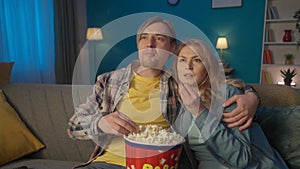 A couple is watching a movie, eating popcorn. Man and woman hugging, relaxing on a sofa in the living room, enjoying a