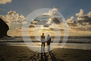 Couple watching sunset at Piha Beach, sunbeams shining through the clouds. Waitakere, Auckland