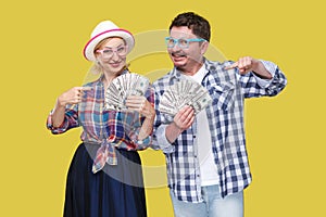 Couple of wealthy friends, adult man and woman in casual checkered shirt standing together holding fan of dollars and pointing