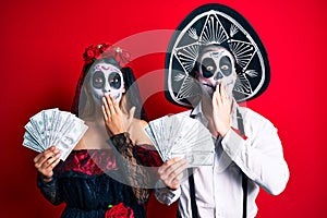 Couple wearing day of the dead costume holding dollars covering mouth with hand, shocked and afraid for mistake