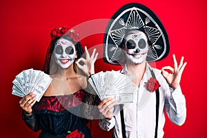 Couple wearing day of the dead costume holding dollars doing ok sign with fingers, smiling friendly gesturing excellent symbol