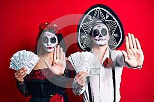 Couple wearing day of the dead costume holding dollars with open hand doing stop sign with serious and confident expression,