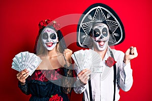 Couple wearing day of the dead costume holding dollars screaming proud, celebrating victory and success very excited with raised