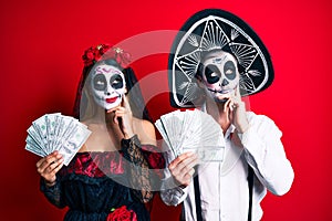 Couple wearing day of the dead costume holding dollars serious face thinking about question with hand on chin, thoughtful about