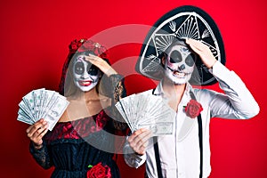 Couple wearing day of the dead costume holding dollars stressed and frustrated with hand on head, surprised and angry face