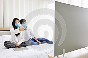 Couple wearing facial mask watching news on television while staying at home during quarantine from coronavius or covid-19