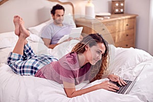 Couple Wearing Pyjamas Lying In Bed At Home With Woman Using Laptop And Man Digital Tablet