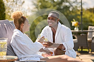 Couple Wearing Robes Outdoors Sitting With Drinks On Loungers Around Swimming Pool On Spa Day