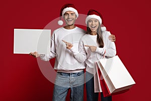 Couple Wearing Santa Hats Holding Shopping Bags And Pointing At Blank Placard