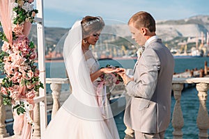 Couple in wedding arch exchange rings with lake on background, the bride with long beautiful hairs and groom in black