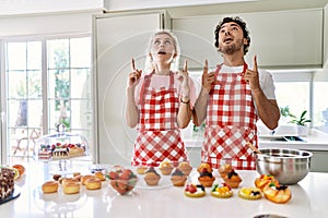 Couple of wife and husband cooking pastries at the kitchen amazed and surprised looking up and pointing with fingers and raised