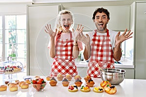 Couple of wife and husband cooking pastries at the kitchen celebrating crazy and amazed for success with arms raised and open eyes