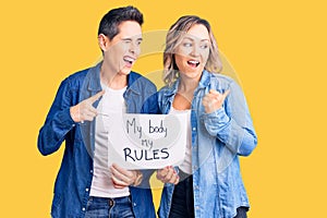 Couple of women holding my body my rules banner pointing thumb up to the side smiling happy with open mouth