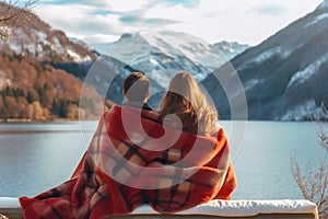 A couple wrapped in a blanket sits intimately, admiring the serene view of a mountain lake