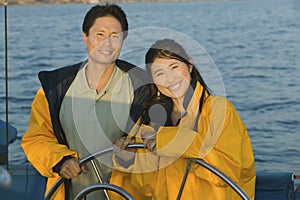 Couple In Yellow Anoraks At Steering Wheel Of Sailboat