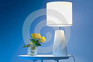 A couple of yellow roses on a white table next to a lamp against dark blue background close up floral view