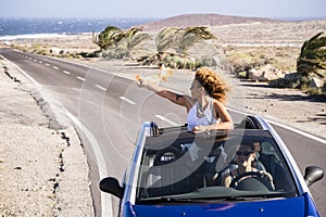 Couple of young adult women travel together in blue convertible car in a long asphalt road with ocean in background - concept of