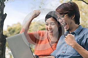 Couple young asian man and woman working with laptop and enjoying cheerful his online winner success  on a bench in the park