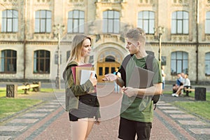 Couple of young attractive students talking outdoors in a park near the university buildong holding laptop, books and notebooks.