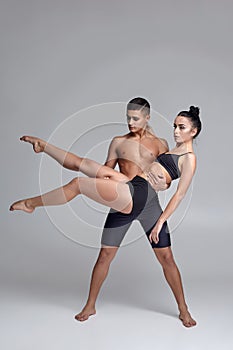 The couple of a young modern ballet dancers in black suits are posing over a gray studio background.