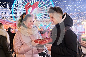 Couple of young people at Christmas market. Happy boy and girl giving gift box