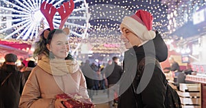 Couple of young people at Christmas market. Happy holiday boy and girl giving gift box
