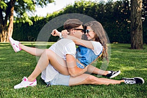 Couple of young people on grass in summer park. Pretty girl with long curly hair in shorts is sitting on handsome guy in